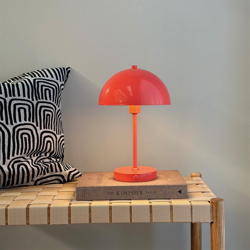 Rød bordlampe står tændt på en bog (THE KINFOLK TABLE) på en flettet bænk; sort-hvid geometrisk pude til venstre, neutral væg i baggrunden.