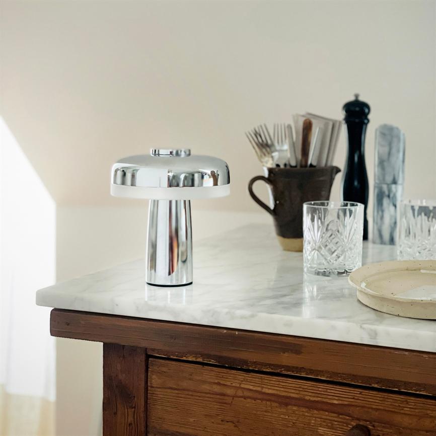 En krom bordlampe står på en hvid marmorplade; i baggrunden en krukke med bestik, en peberkværn, krystalglas og en rund tallerken på et træskab.