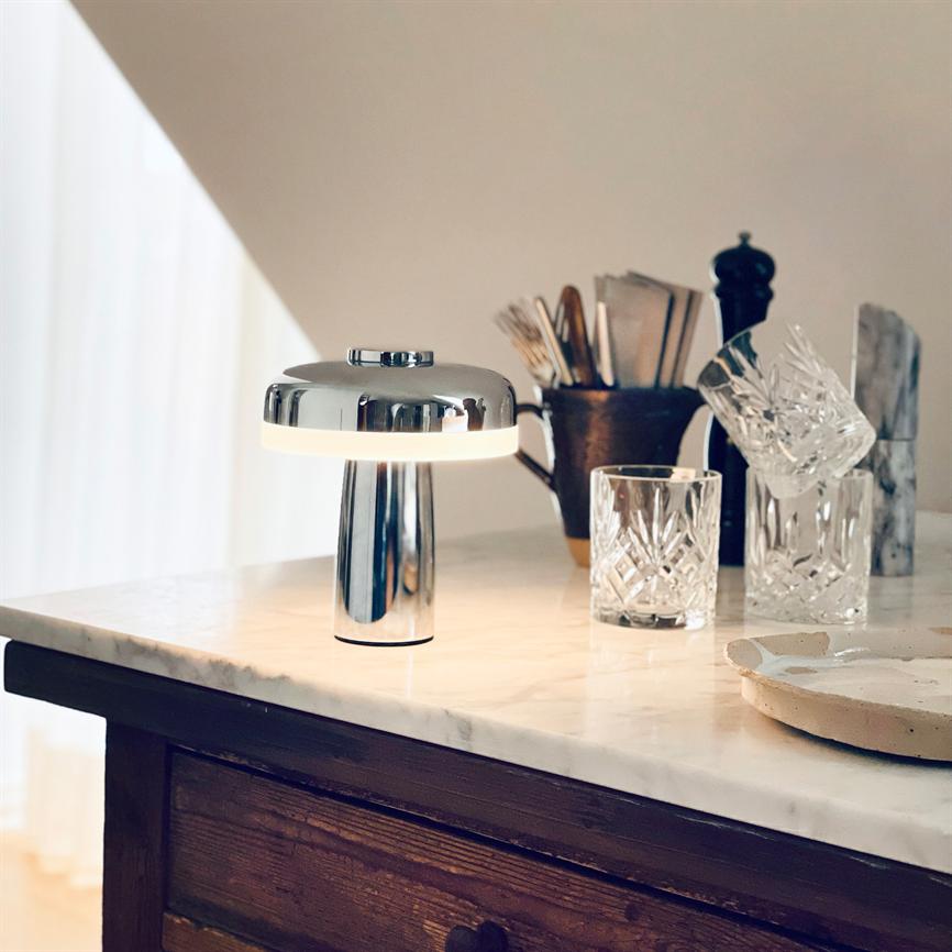 Bordlampe i krom sender blødt lys på en marmorbordplade, omgivet af krystalglas, et bestikkrus og et serveringsfad på et mørkt træskab i et lyst køkkenhjørne.