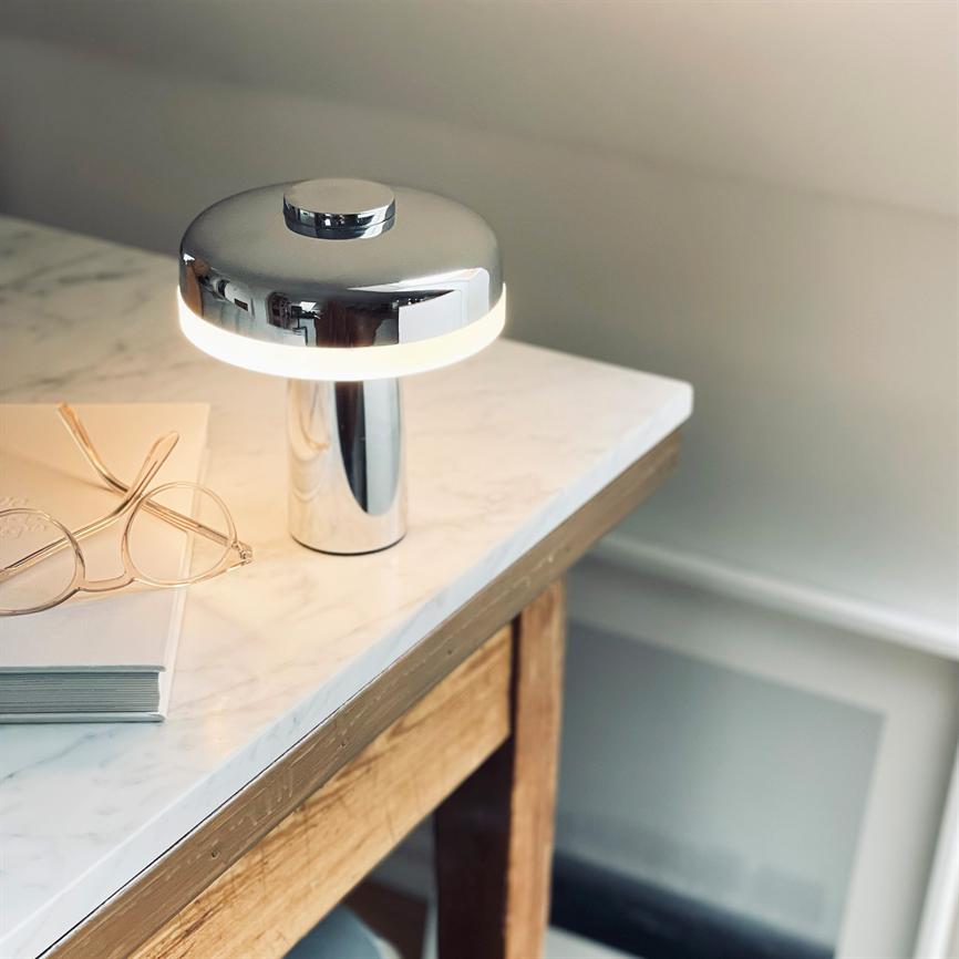Sølv bordlampe udsender blødt, varmt lys på en hvid marmoreret bordplade; et par gennemsigtige briller ligger ovenpå en bog på et træbord tæt ved væggen.