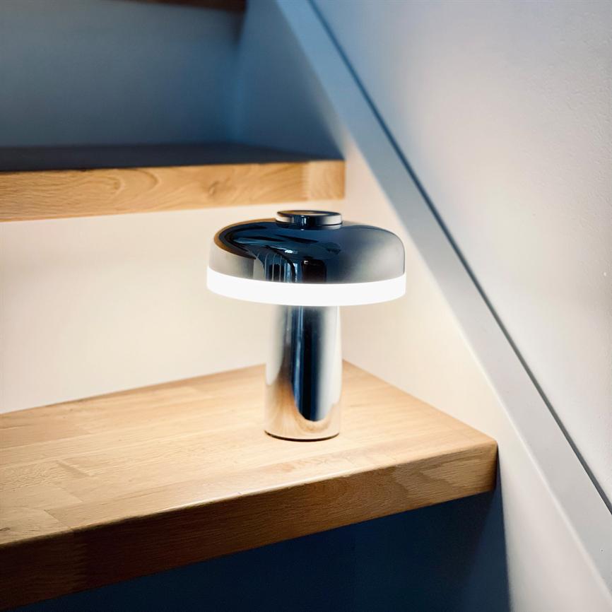 En moderne, svampeformet bordlampe udstråler blødt hvidt lys og står på et lyst trappetrin af træ under en skrå, hvid væg.