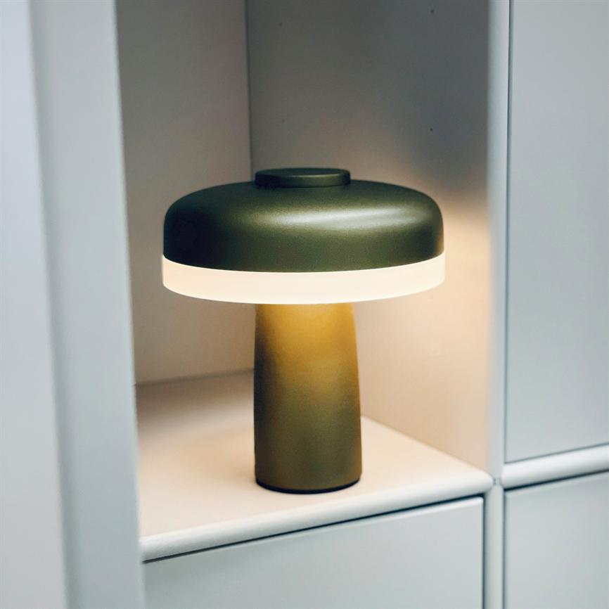 En olivengrøn bordlampe lyser blødt fra sin hvide diffuser, stående på en hylde i en hvid indbygningsreol, hvilket skaber et varmt, afdæmpet lys i nichen.