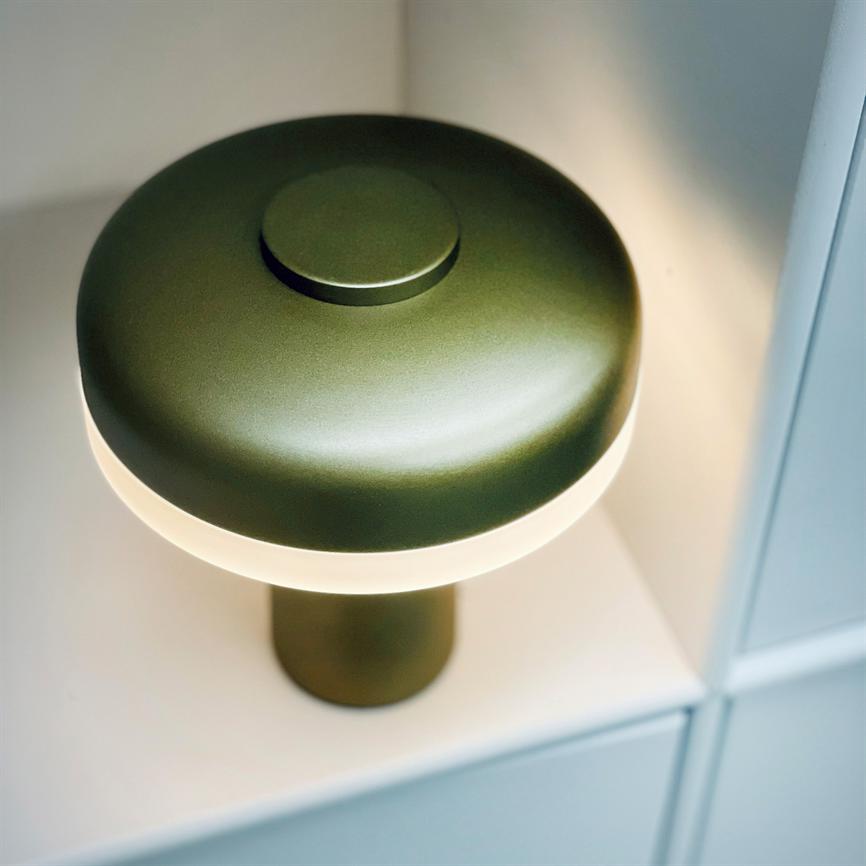 En grøn, svampeformet bordlampe med mat finish udsender blødt, varmt lys; den står på en hvid hylde i et modulopbygget reolsystem.
