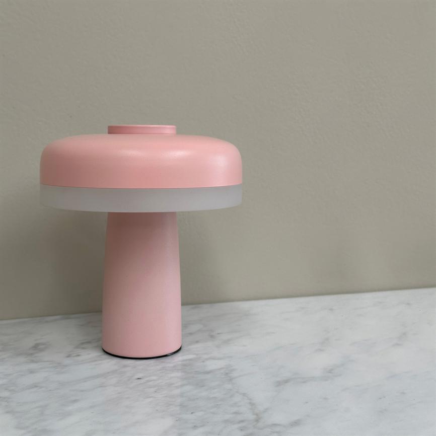 Lyserød svampeformet bordlampe står på et hvidt marmorbord foran en ensfarvet beige væg; lampen har en mat hvid diffusionsring og en glat, cylindrisk fod.