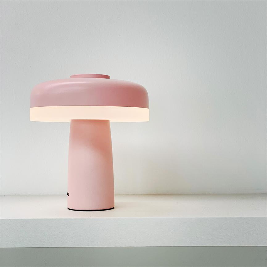 Rosa, svampeformet bordlampe står tændt på en hvid hylde og udsender blødt, varmt lys mod en lys, neutral væg.