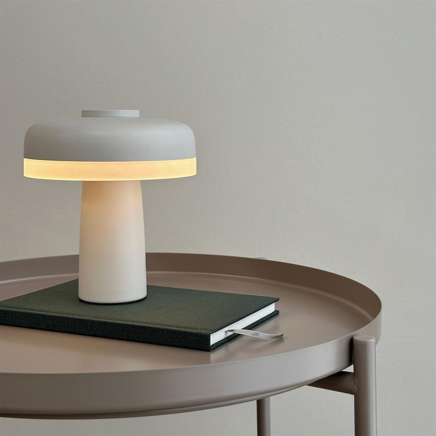 Lampe udsender blødt, varmt lys; står på en grøn indbundet notesbog på et rundt, beige metal-bord foran en lys grå væg.