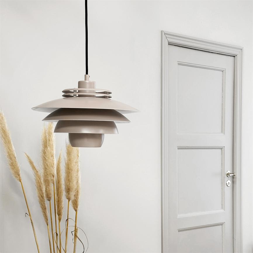 En lagdelt, beige pendellampe hænger i sort ledning, midt for en hvid væg; pampasgræs står til venstre, og en lukket dør med paneler ses til højre.