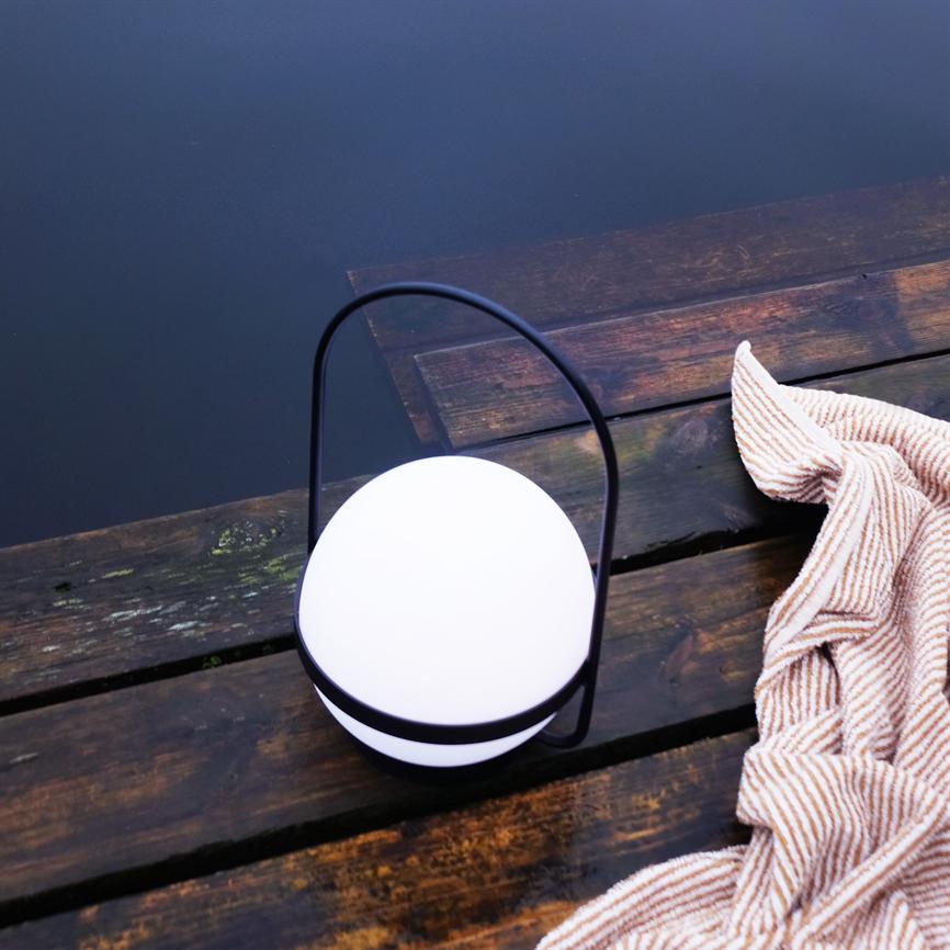 En rund bærbar lampe med sort håndtag lyser blødt hvidt og står på et vådt, mørkt trædæk ved en stille sø; et foldet, stribet tæppe ligger ved siden af.