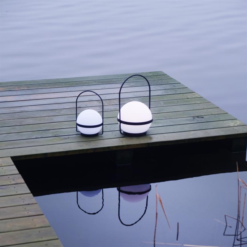 To runde, hvide lanterner står på kanten af en træbro; de udsender blødt lys, spejler sig i det stille søvand og ligger i et roligt, blågråt miljø.