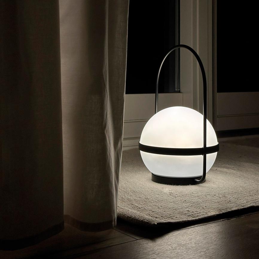 En kugleformet bordlampe med sort bøjle udsender blødt hvidt lys og står på et lille tæppe foran en lukket glasdør om natten, delvist skjult af et gardin.