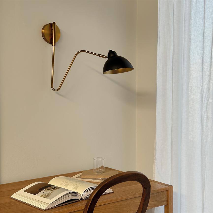 Væglampe med sort skærm og gylden arm strækker sig over et træskrivebord, hvor en åben bog, et par bøger og et tomt glas ligger, ved siden af et lyst, hvidt gardin.