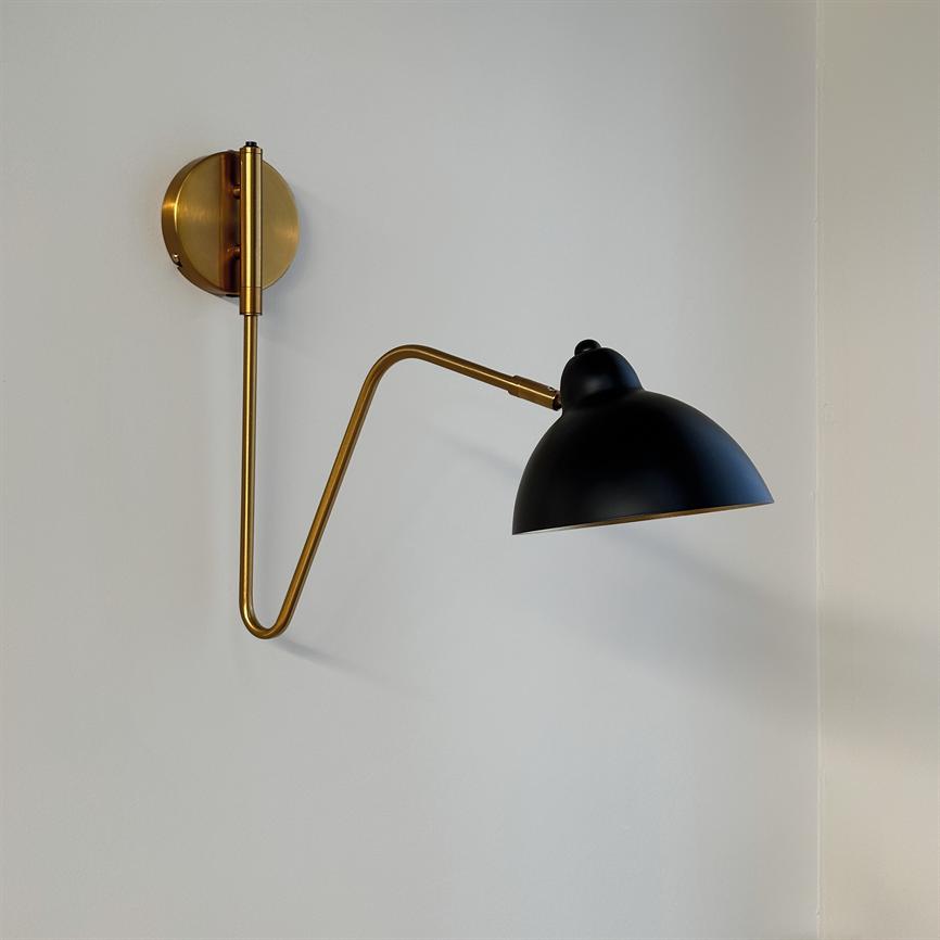 Væglampe: en messing vægmonteret arm strækker sig i en zigzag og holder en mat sort halvkugleformet skærm mod en lys, neutral væg i et hjørne.