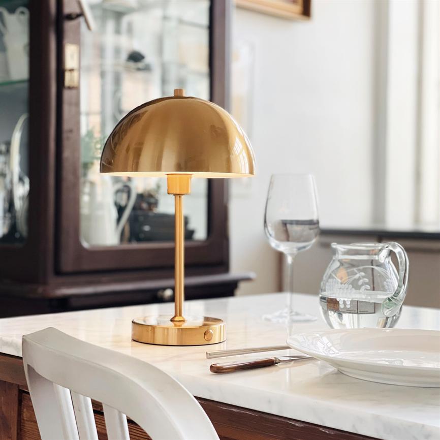 Guldfarvet bordlampe står tændt på et hvidt marmorbord; kaster blødt lys over en dækket plads med vinglas, vandkaraffel, kniv og tallerken; vitrineskab og stol i baggrunden.
