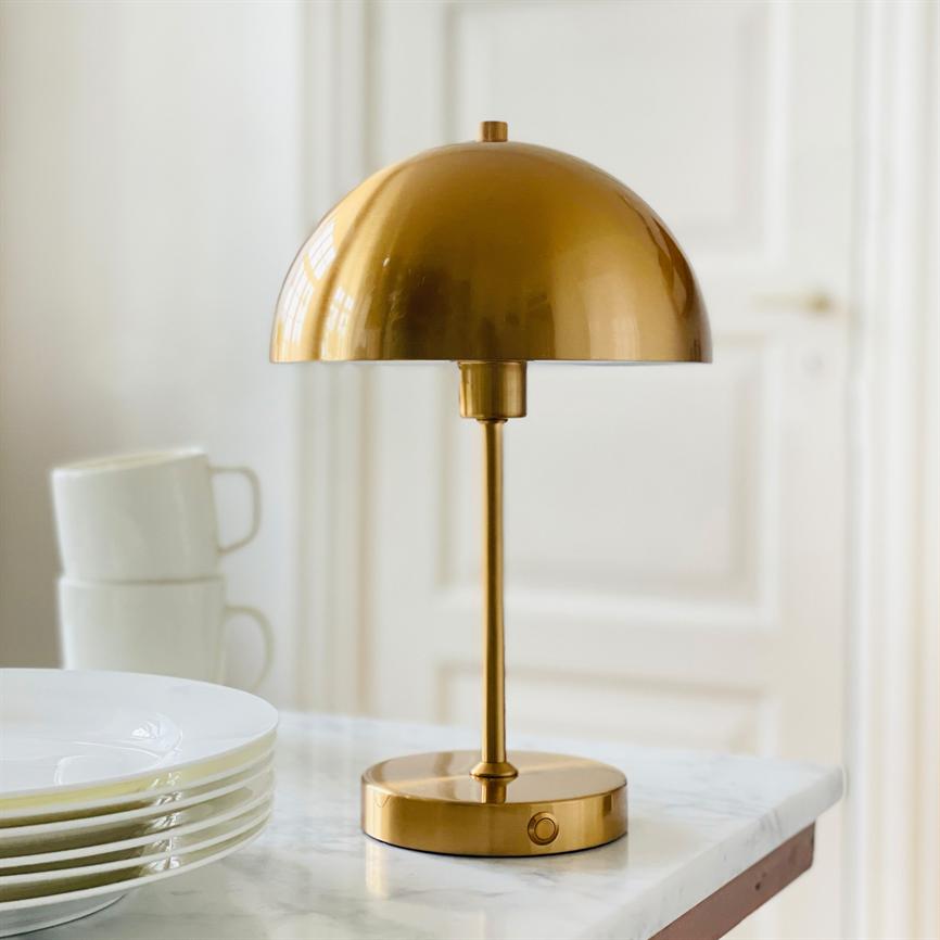 Guldbordlampe står på en hvid marmorbordplade; den slanke stang og kuppelformet skærm reflekterer lys. Ved siden af ligger en stak hvide tallerkener og to kopper, hvid dør bagved.