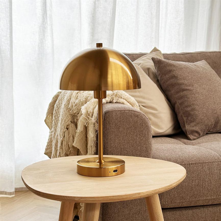 Guldfarvet bordlampe står på et rundt lyst træbord og kaster blødt lys i et stuehjørne med beige sofa, flere puder og et krøllet lyst tæppe foran let gennemsigtige hvide gardiner.