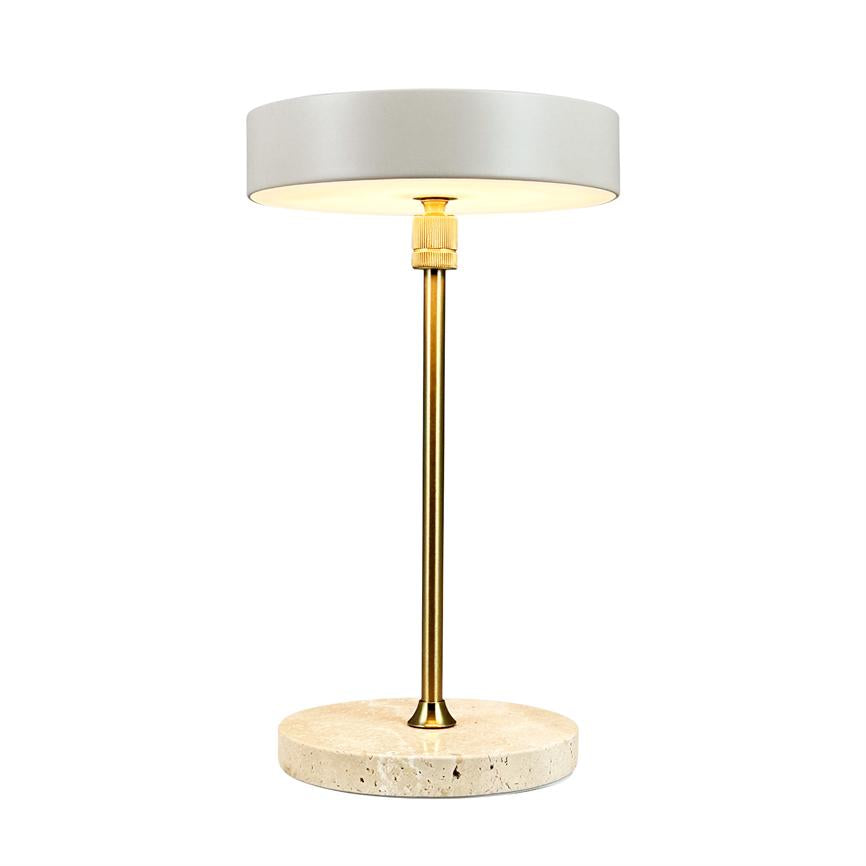 Bordlampe står tændt; hvid cylindrisk skærm afgiver varmt lys, monteret på en slank messingstang og en rund, lys travertinfod, placeret mod neutral hvid baggrund.