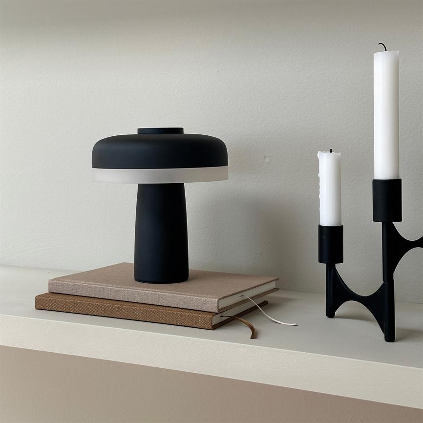 Sort bordlampe står på to stabler notesbøger, ved siden af en sort lysestage med to slukkede hvide stearinlys, på en lys cremefarvet hylde foran en neutral væg.