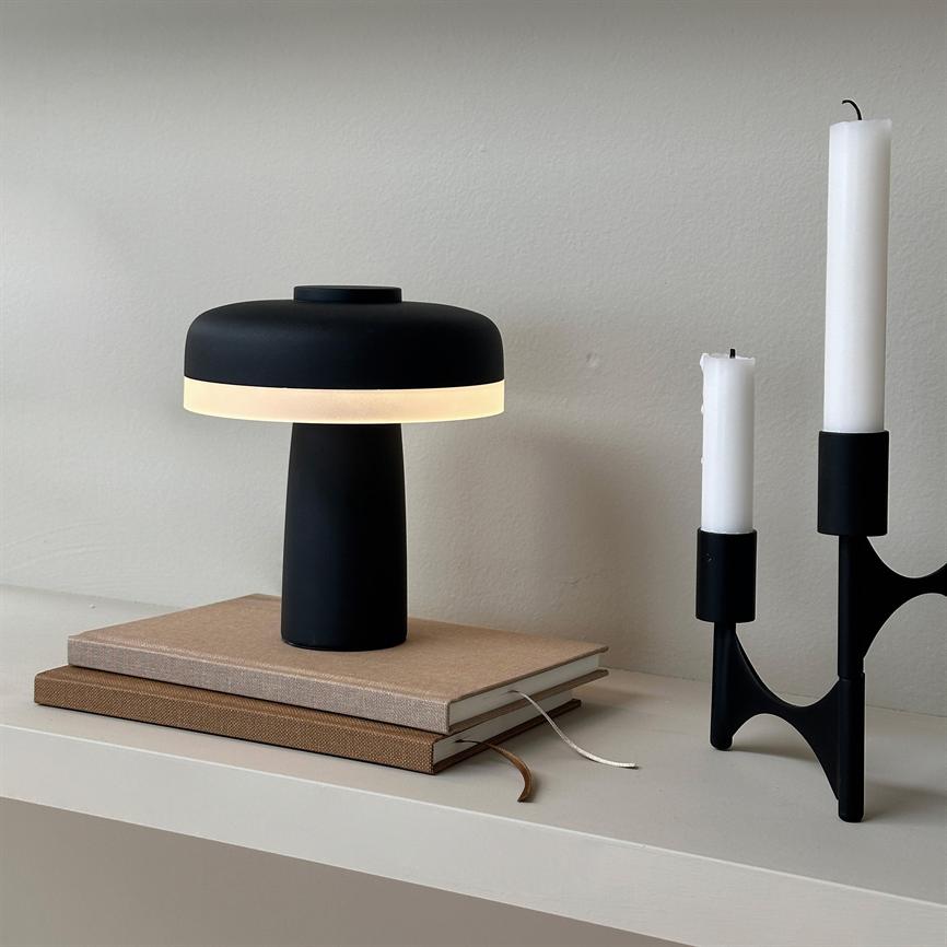 Lampe lyser blødt; står på to stabler notesbøger på en hvid hylde, ved siden af en sort lysestage med tre hvide stearinlys.
