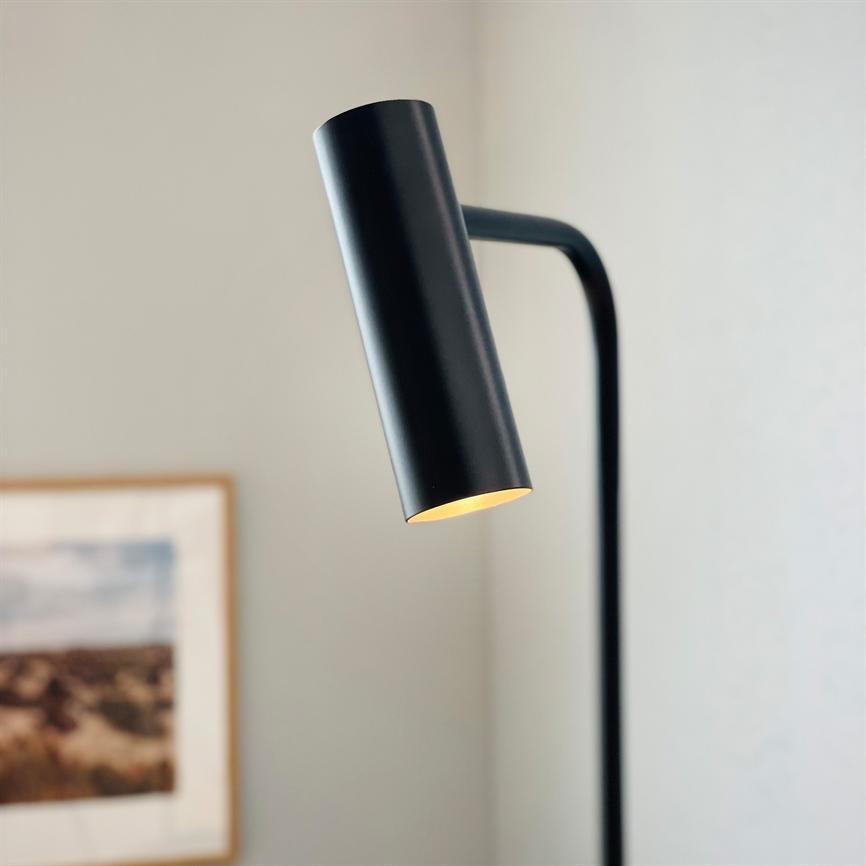 Sort, cylinderformet gulvlampe lyser svagt nedad fra en buet stang i et lyst, minimalistisk værelse med en indrammet landskabsfotografi på væggen.