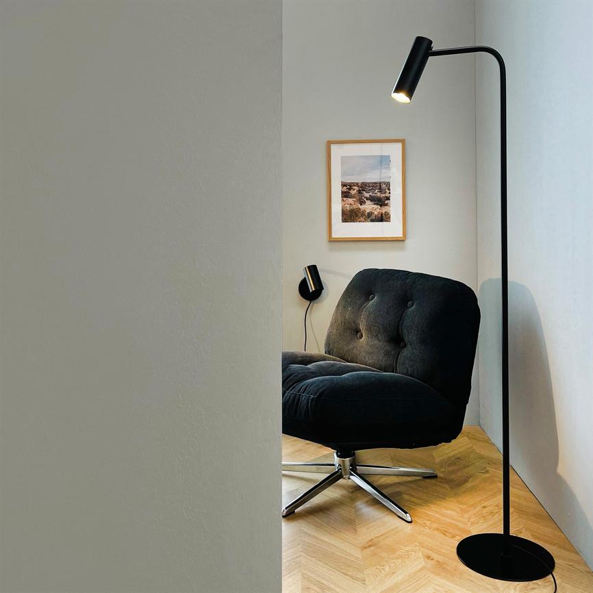 En mørkgrå, knappet lænestol står i et hjørne, oplyst af en sort gulvlampe; parketgulv, hvid væg og et indrammet landskabsfoto i baggrunden.