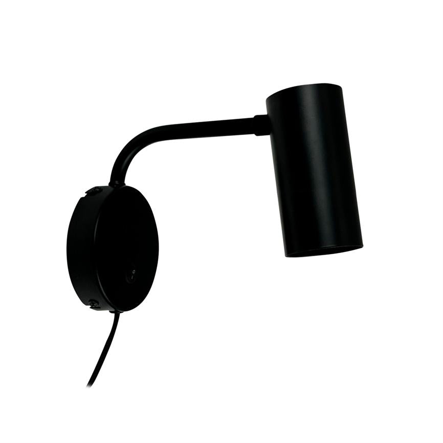Sort metal væglampe monteret på rund vægplade, arm bøjet udad og cylinderformet lampehoved peger skråt nedad; enkel, moderne design mod hvid baggrund.
