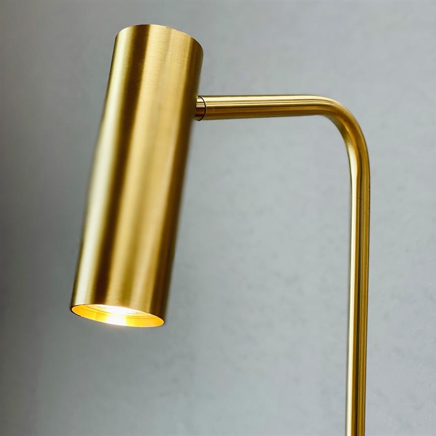 Messingfarvet bordlampe med cylindrisk skærm, tændt og kaster varmt lys nedad; arm bøjet i en ret vinkel foran en lys grå væg.