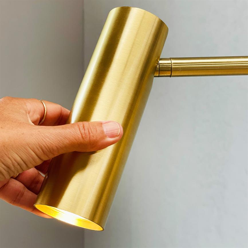 En cylindrisk, messingbelagt lampe holdes og justeres af en hånd; lyset skinner fra lampens åbning, monteret på en tynd arm foran en lys grå væg.