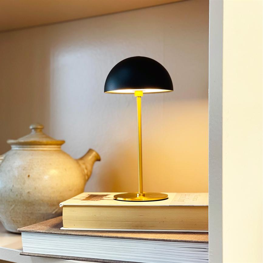 Bordlampe med sort halvkugleskærm lyser varme gule toner; den står på en stak bøger på en hylde ved en rustik keramisk tekande i et blødt, varmt hjørne.