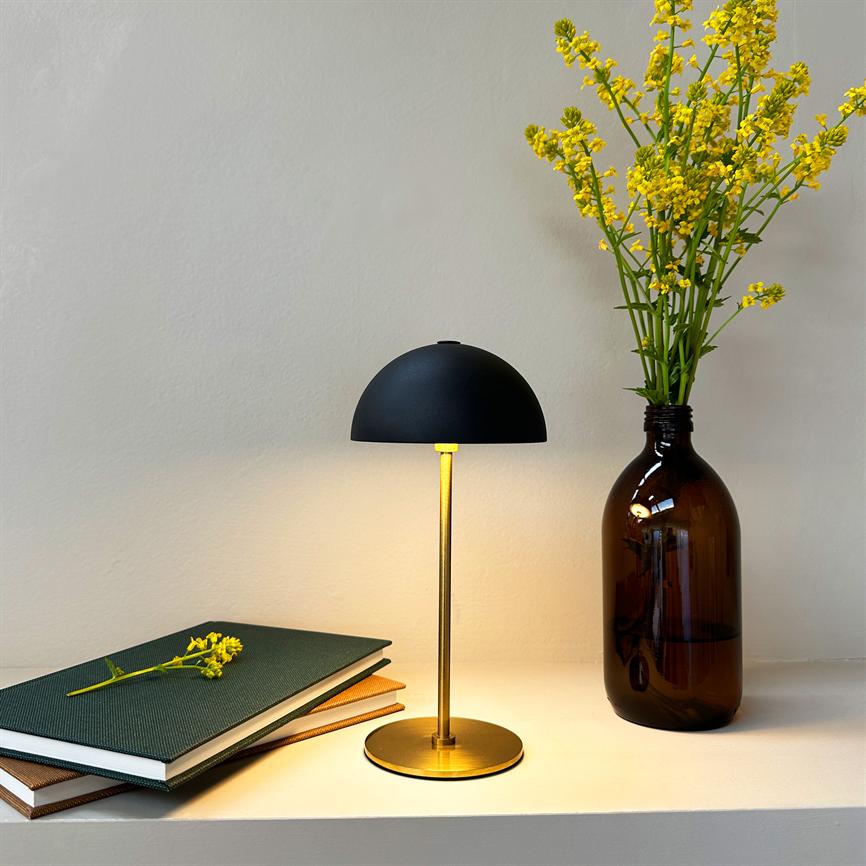 Lampe med sort kuppel på messingfod udsender varmt lys på en hvid hylde; ved siden ligger en stak notesbøger med en blomst og en brun flaskevase med gule blomster.