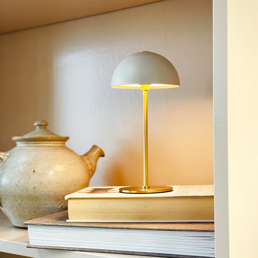 Bordlampe med rund skærm lyser blødt og varmt, stående på en stak bøger i en hylde, med en beige keramisk tekande placeret ved siden af.