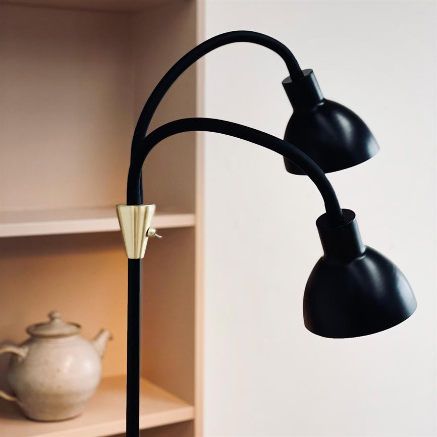 Gulvlampe med to buede arme bærer to sorte metalkærme, placeret foran en lys væg og ved siden af en beige hylde med en keramisk tekande.