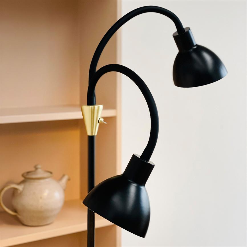 Sort gulvlampe med to buede arme og matte skærme, med en messingdetalje og lille vippekontakt; den står slukket ved en lys træreol med en beige tekande på hylden.