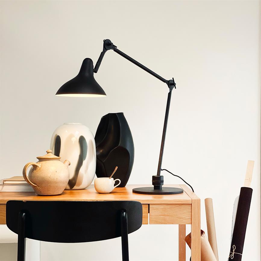 En sort justerbar skrivebordslampe lyser over et træbord; den oplyser keramiske vaser, en tekande og en kop med ske, mens en sort stol og rullepapirer står i et lyst, minimalistisk rum.