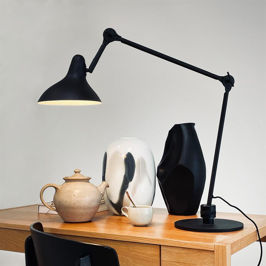 Sort, justerbar skrivebordslampe kaster varmt lys over en lertekande og en kop på et lyst træskrivebord, flankeret af en hvid og en sort keramisk vase foran en neutral væg.