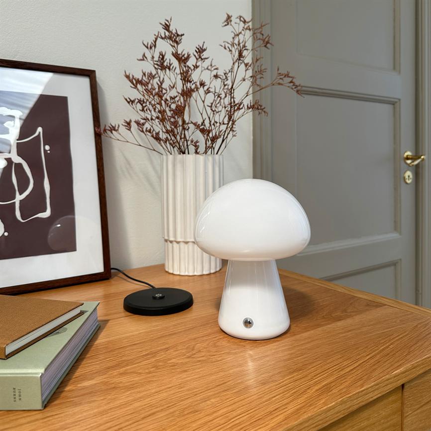 Hvid svampeformet lampe står slukket på et træskab, ved en vase med tørrede grene og indrammet abstrakt kunst, foran en lukket hvid dør.