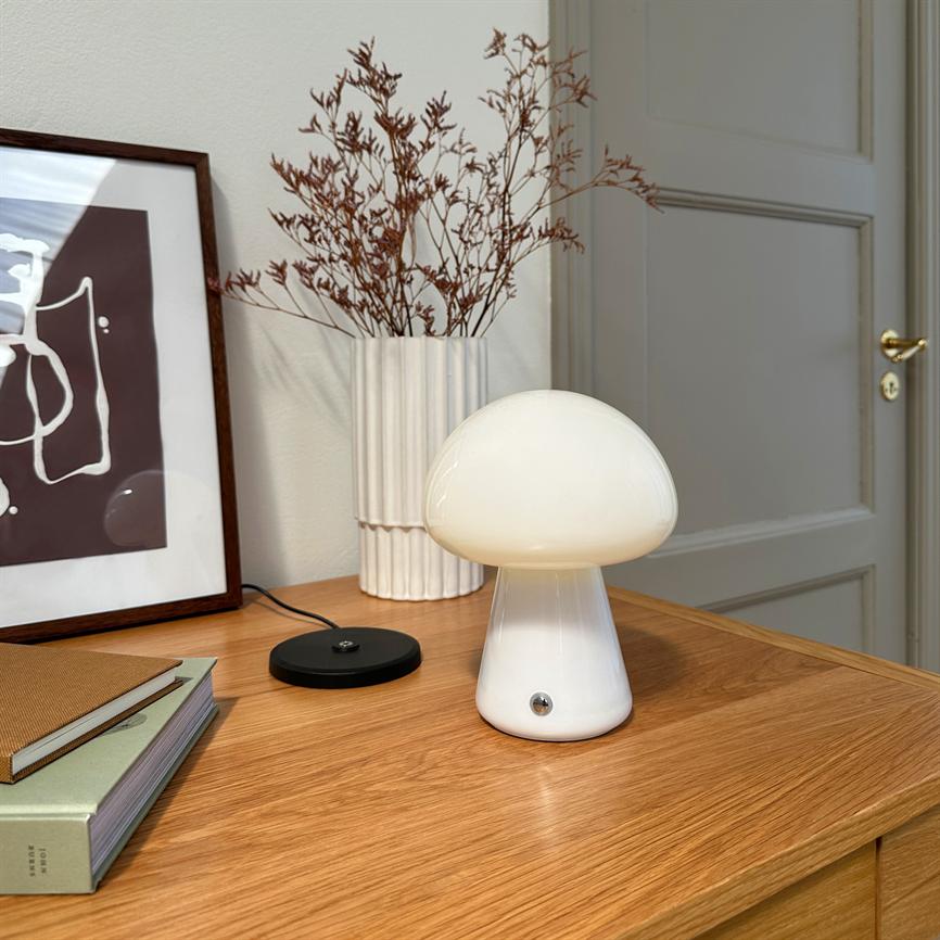 En hvid svampeformet bordlampe lyser blødt og står på et træskab ved en hvid keramikvase med tørrede grene, et indrammet abstrakt billede, nogle bøger og en lukket dør.
