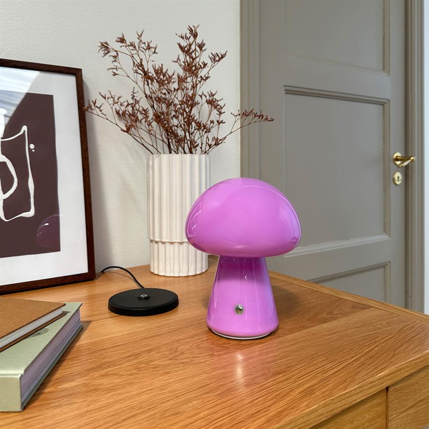 Lyserød svampeformet bordlampe står på et træskab, omgivet af en hvid rillede vase med tørrede grene, en indrammet plakat og en lukket dør i baggrunden.