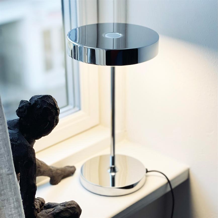 En krom bordlampe kaster blødt lys på en hvid vindueskarm ved et vindue; ved siden af står en mørk, siddende skulptur, med et lyst gardin og hvid væg i baggrunden.