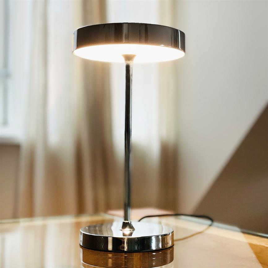 Metalbordlampe lyser varmt nedad; tynd stang forbinder rund skærm og rund, reflekterende base på et glasbord med slørede gardiner og et vindue i baggrunden.