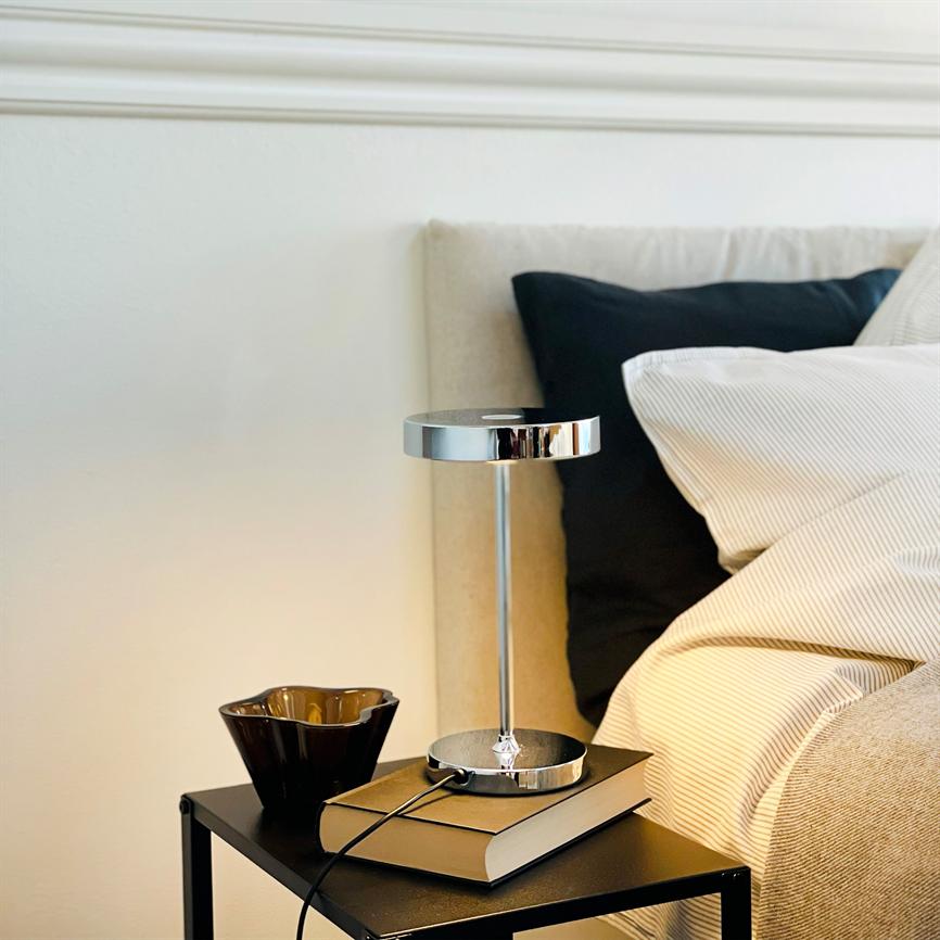 Bordlampe står på en bog på et lille sort sengebord; lampens chrome reflekterer lyset. En brun keramikkop ved siden af, beige sengegavl, stribet hvidt sengetøj og sort pude i baggrunden.