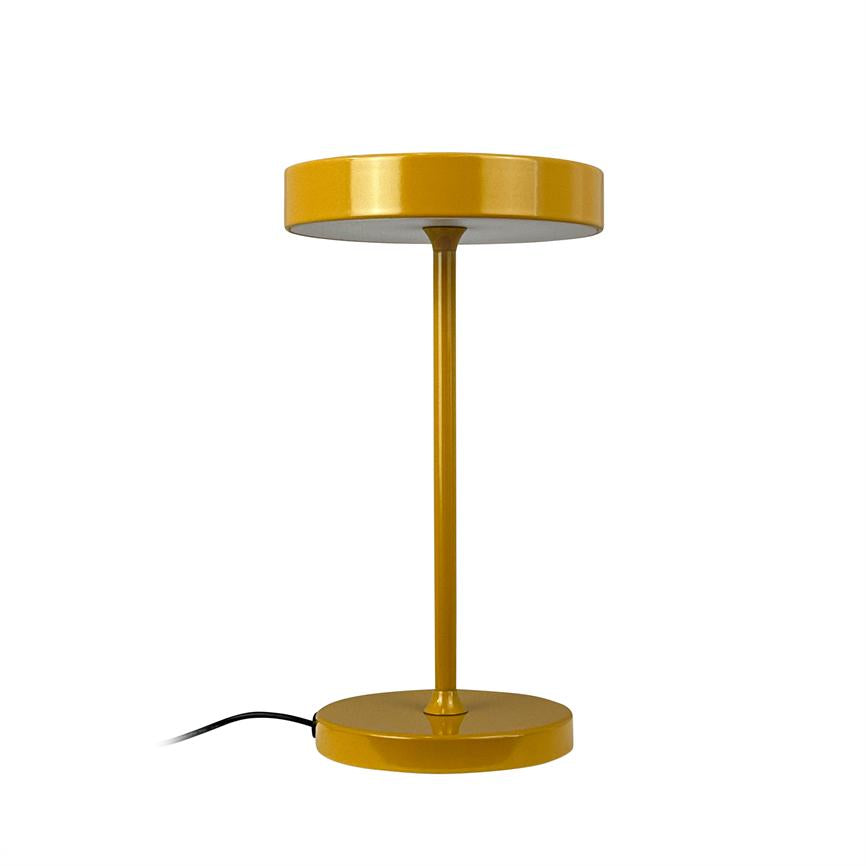 Gul metalbordlampe står på hvid baggrund; en slank stang forbinder rund base og lavprofilskærm, og en sort ledning strækker sig svagt ud til siden.