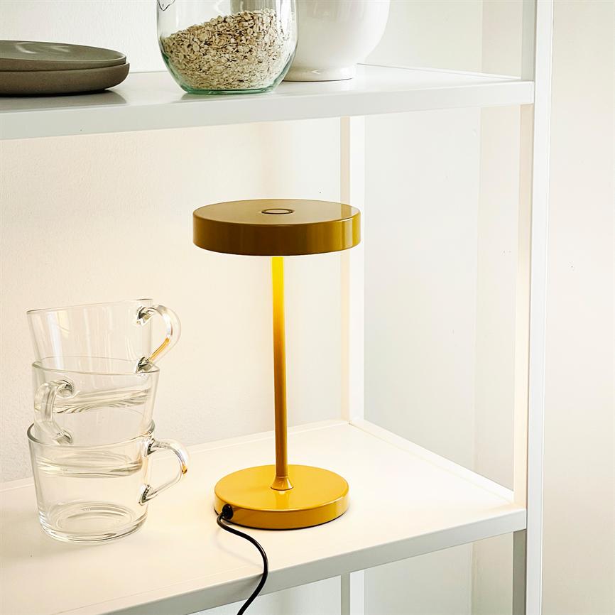 Gul bordlampe står tændt på en hvid hylde; dens sorte ledning slynger sig mod kanten; tre gennemsigtige kopper er stablet ved siden af, og øverste hylde rummer glas- og porcelænsbeholdere.