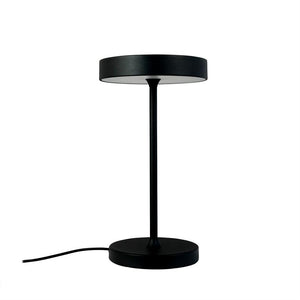 Sort, moderne bordlampe står oprejst med tynd stang og cirkulært skærm; rund fod med synlig strømkabel mod venstre på hvid baggrund.