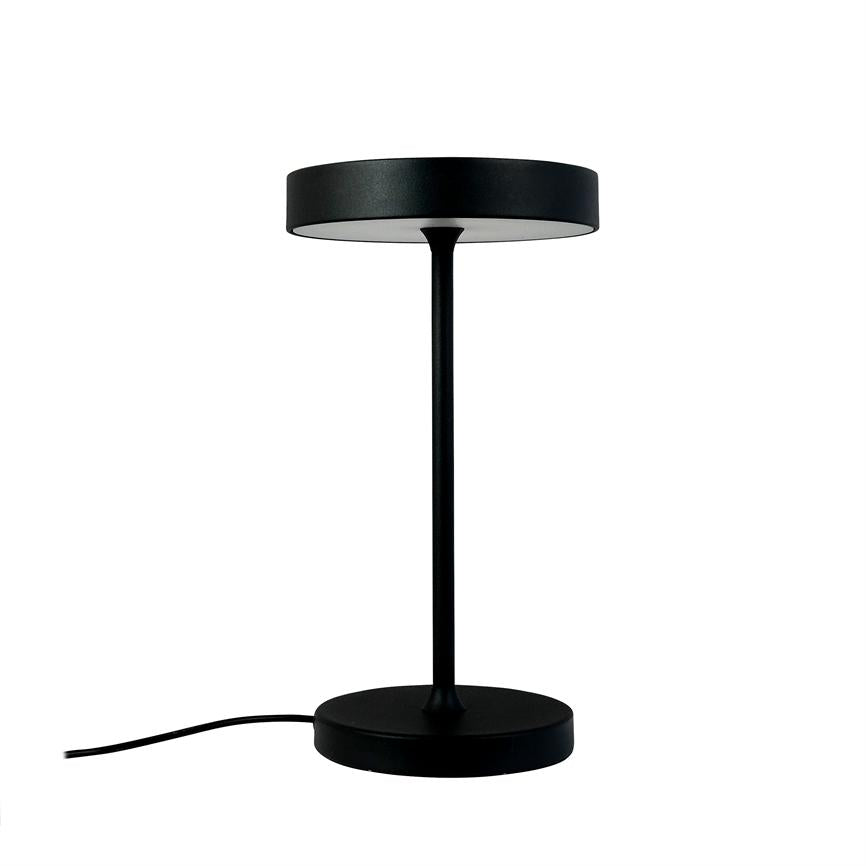 Sort, moderne bordlampe står oprejst med tynd stang og cirkulært skærm; rund fod med synlig strømkabel mod venstre på hvid baggrund.
