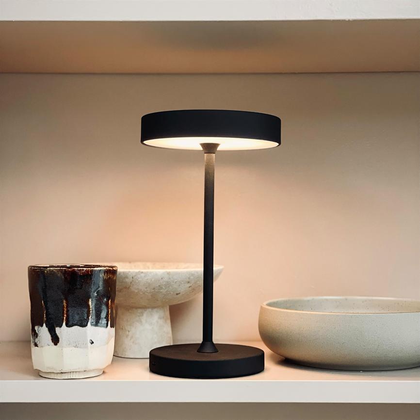 Slank sort bordlampe står tændt på en hylde, kaster blødt, varmt lys; omgivelsen består af keramikkrus og skåle i neutrale toner på en hvid baggrund.