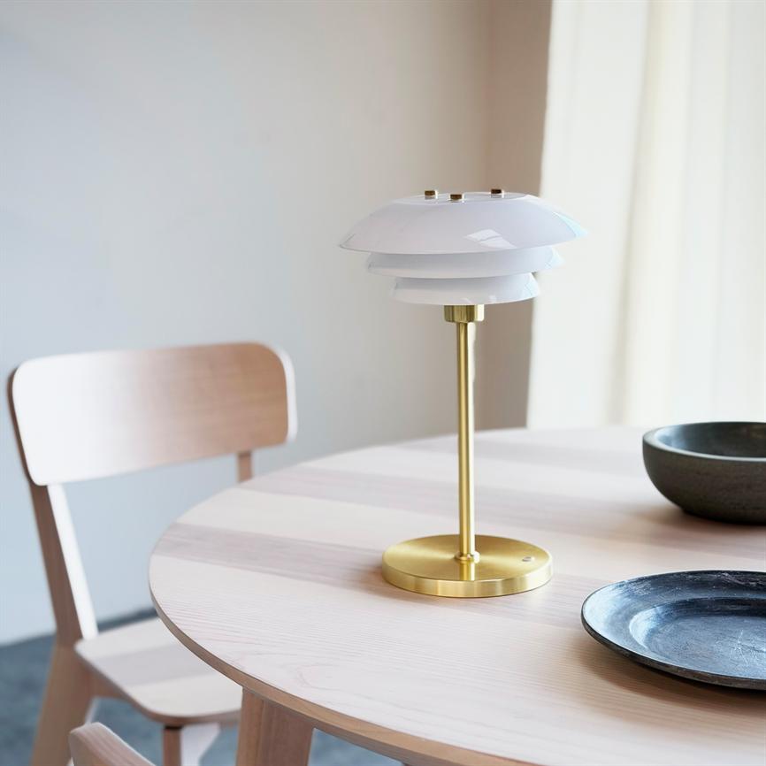 Lampe med hvid, lagdelt skærm og messingfod står på rundt lyst træbord i et lyst, minimalistisk spisehjørne med træstol og keramiske skåle.