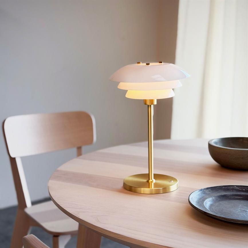 Bordlampe med gylden metalfod og lagdelt hvid skærm står tændt på et rundt lyst egetræsbord, kaster varmt lys; en enkel træstol og to mørke skåle i baggrunden.