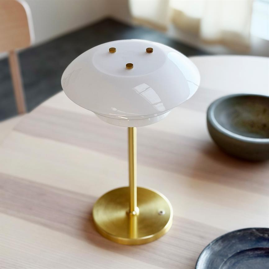 Bordlampe i messing med rund hvid glasskærm står på et lyst træbord; nær en grå keramisk skål og en sort tallerken i et lyst, diffus oplyst rum.