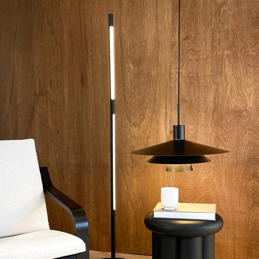 En slank stående LED-gulvlampe oplyser et hjørne med en sort pendellampe over et rundt sort sidebord, hvor en bog og et hvidt krus står foran en træbeklædt væg.