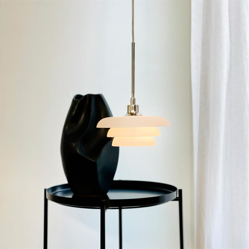 En lagdelt pendel hænger og udsender varmt lys over et rundt, sort sidebord; på bordet står en sort skulpturel vase, med en lys, neutral væg og hvidt gardin i baggrunden.